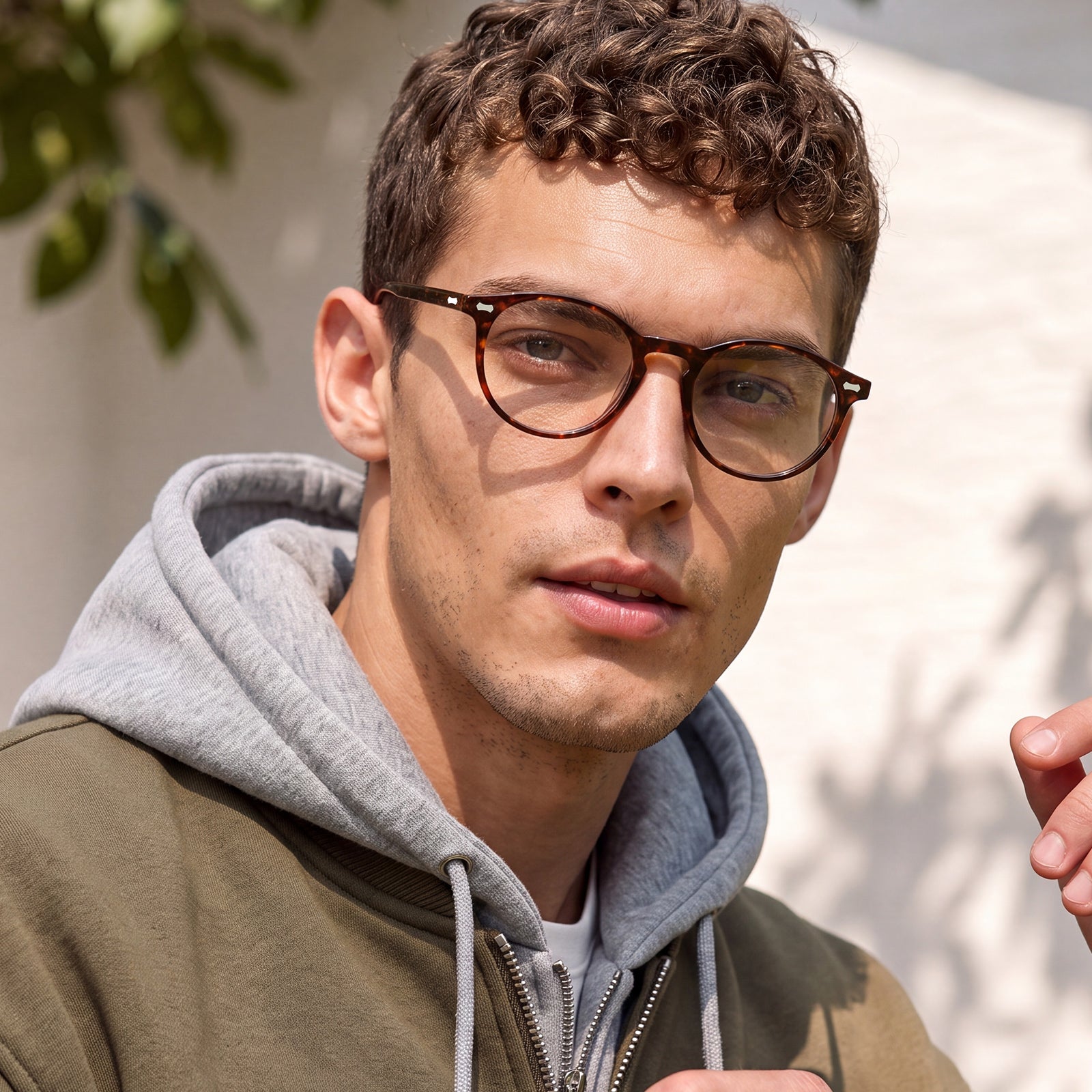 A close-up of a man wearing round brown acetate eyeglasses with a thick browline frame, styled with a gray hoodie and olive green jacket against a softly lit outdoor background.