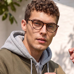 A close-up of a man wearing round brown acetate eyeglasses with a thick browline frame, styled with a gray hoodie and olive green jacket against a softly lit outdoor background.