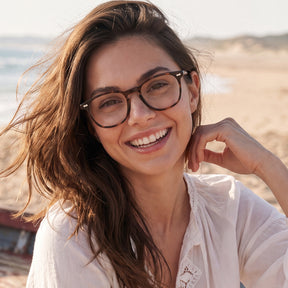 A smiling woman with long brown hair wearing round dark brown eyeglasses, photographed outdoors on a sandy beach with the ocean in the background.