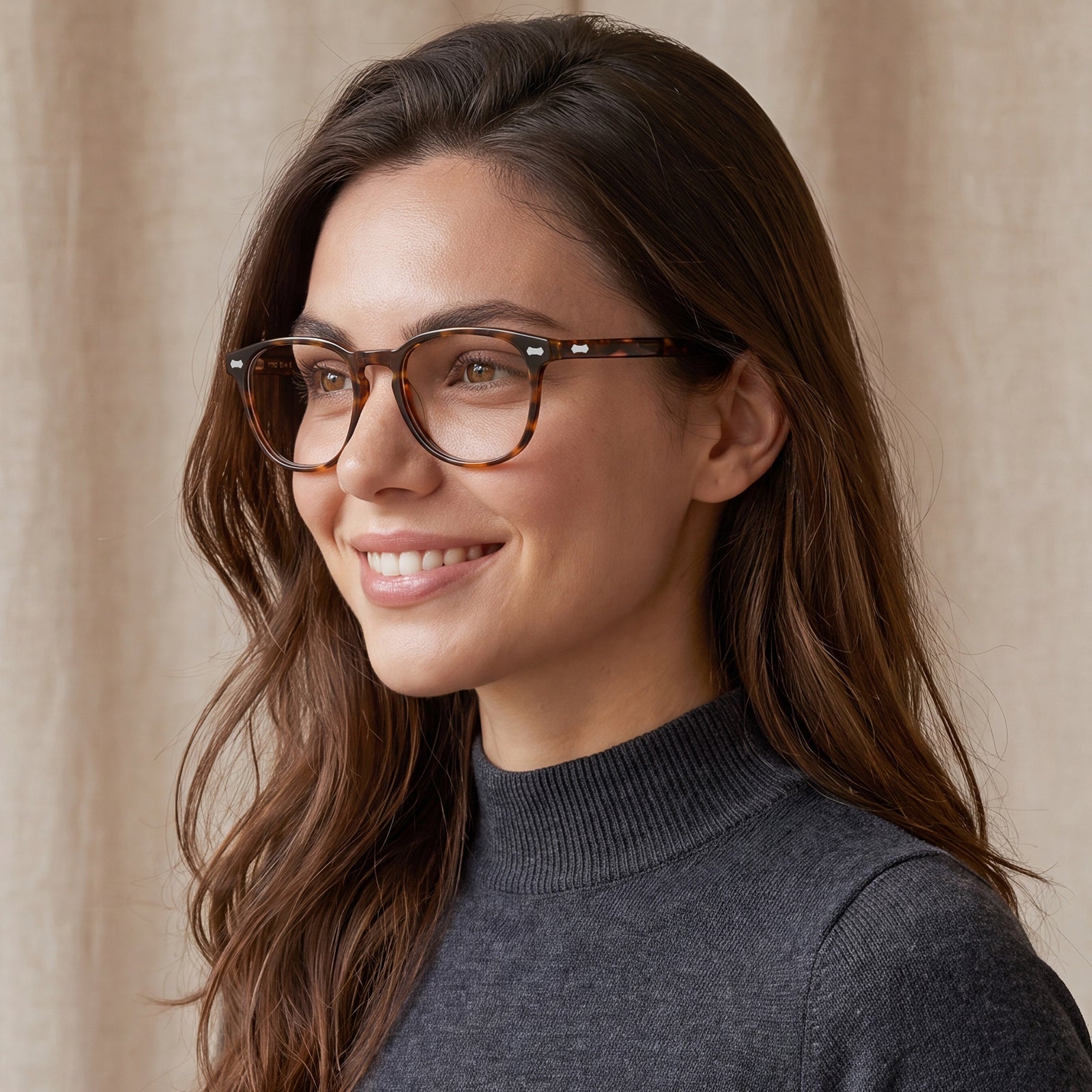 A woman with long brown hair wearing round tortoiseshell eyeglasses and a dark gray ribbed turtleneck sweater, shown in a close-up portrait against a soft beige curtain background.