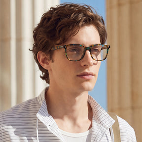 A man wearing tortoiseshell eyeglasses with a square frame, styled with a striped button-up shirt in an outdoor setting 