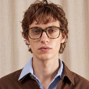 A close-up portrait of a young man with wavy brown hair wearing thick, square tortoiseshell eyeglasses with a green, yellow, and brown pattern, a light blue collared shirt, and a brown jacket