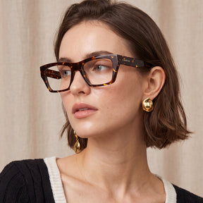 A close-up of a woman wearing rectangular tortoiseshell eyeglasses with the brand name carfia visible on the temple arm, styled with gold earrings against a neutral beige curtain background.