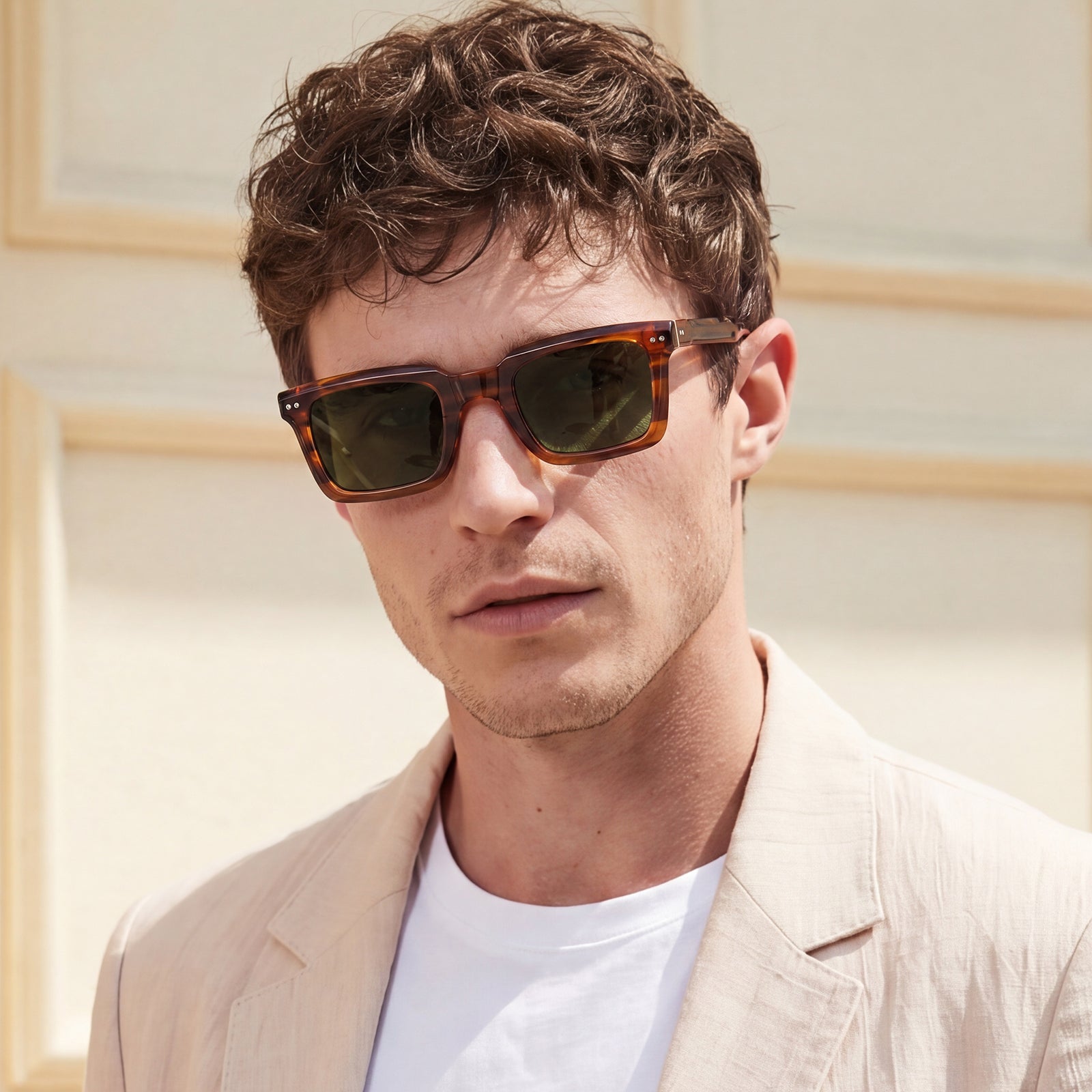 A man wearing tortoiseshell sunglasses with dark lenses and a light beige blazer over a white t-shirt, photographed outdoors against a light beige wall.