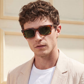 A man wearing tortoiseshell sunglasses with dark lenses and a light beige blazer over a white t-shirt, photographed outdoors against a light beige wall.