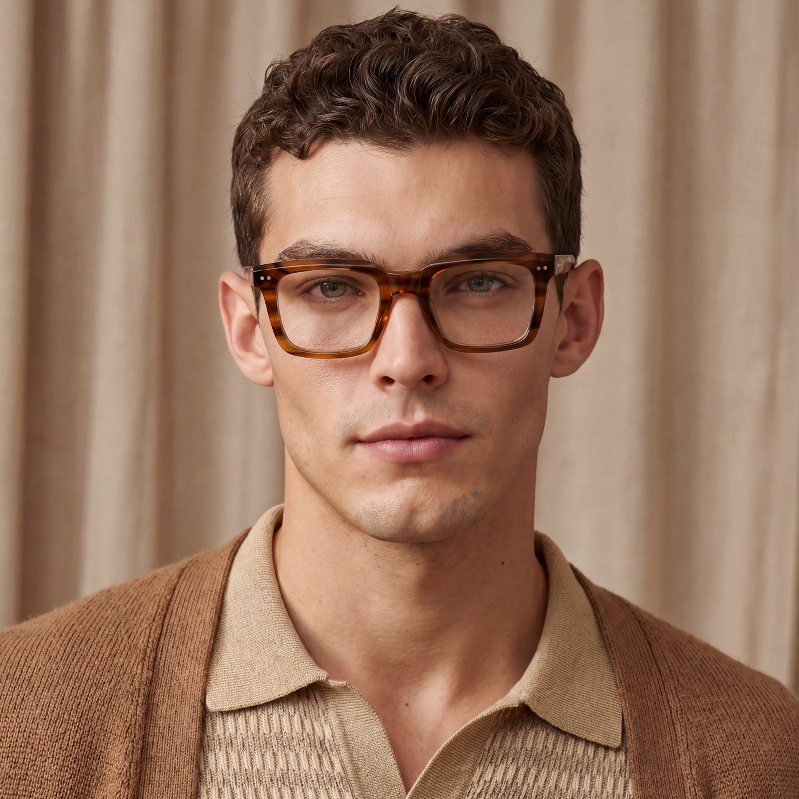 A close-up portrait of a man wearing thick, square tortoiseshell acetate eyeglasses with a warm brown and amber tone, styled with a beige knit polo shirt 