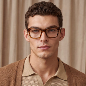 A close-up portrait of a man wearing thick, square tortoiseshell acetate eyeglasses with a warm brown and amber tone, styled with a beige knit polo shirt 