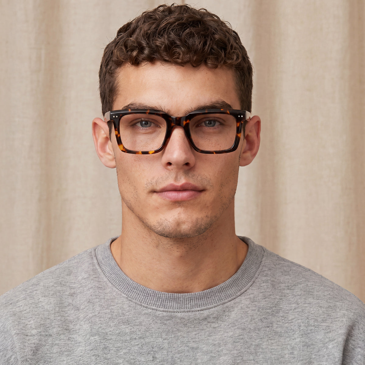 A man wearing tortoiseshell eyeglasses with thick black frames and a gray crewneck sweatshirt 