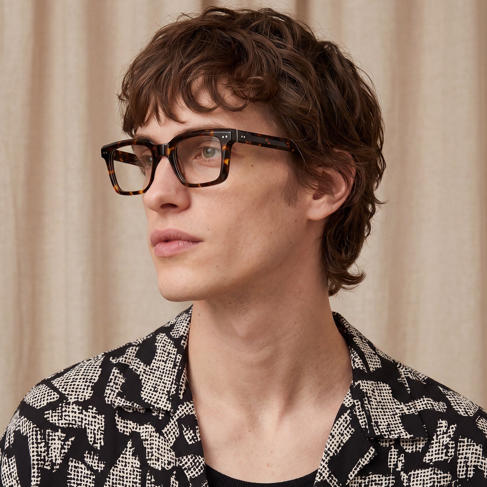 A man with wavy brown hair wearing thick black tortoiseshell eyeglasses and a black-and-white patterned blazer 