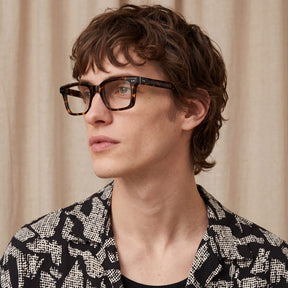 A man with wavy brown hair wearing thick black tortoiseshell eyeglasses and a black-and-white patterned blazer 
