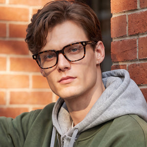 A young man with brown hair and blue eyes wearing tortoiseshell eyeglasses, a gray hoodie, and an olive green sweatshirt, leaning against a red brick wall.