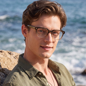 A man outdoors by the ocean wearing rectangular eyeglasses with dark brown frames and an olive green shirt