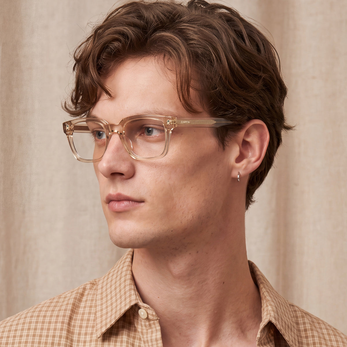 A close-up portrait of a man wearing clear acetate eyeglasses with gold-tone decorative hinges and a tan-and-white checkered shirt, set against a neutral beige curtain background.